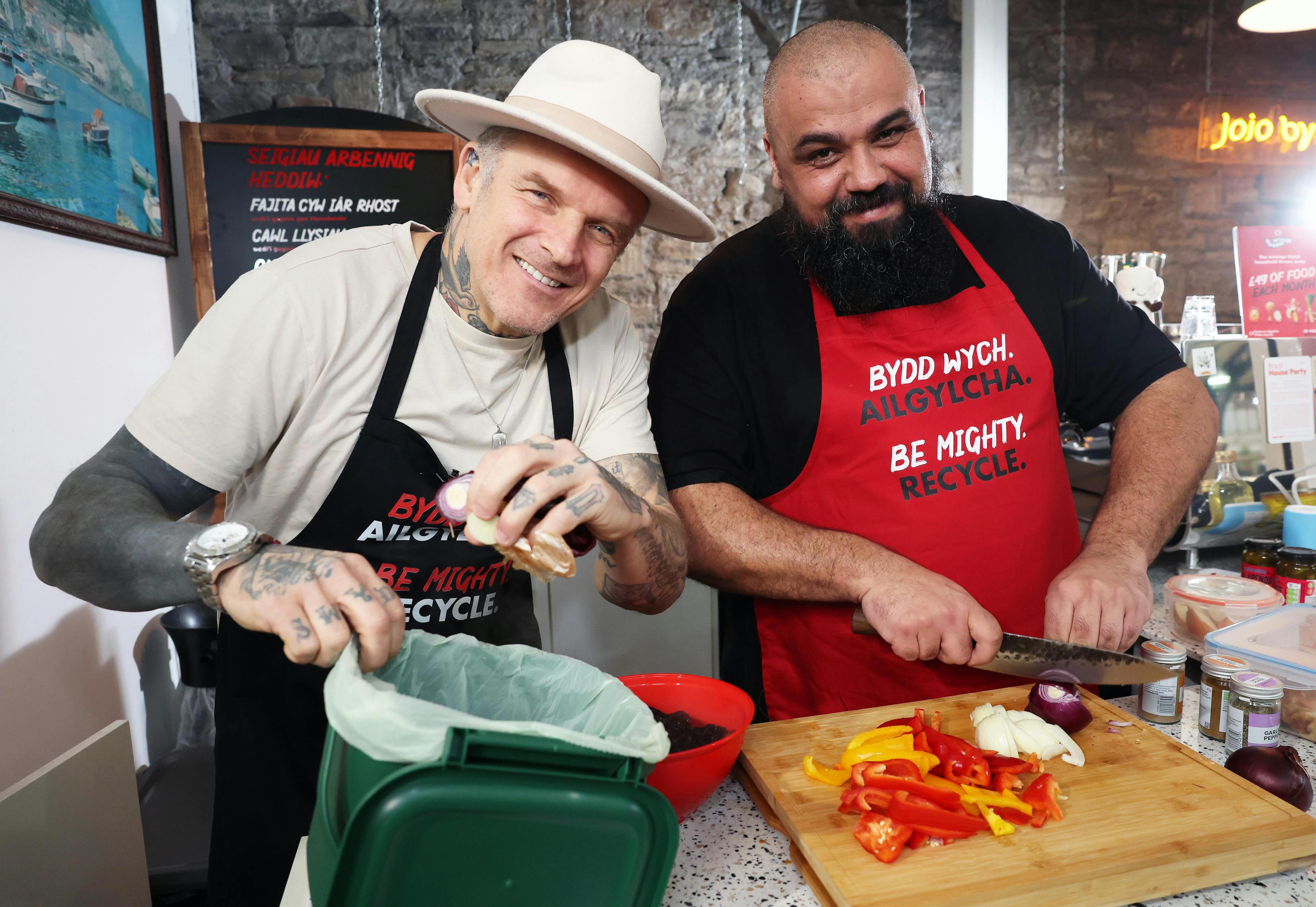 Two chefs in Cardiff Market, one holding a food waste caddy and one chopping vegetables on a wooden chopping board