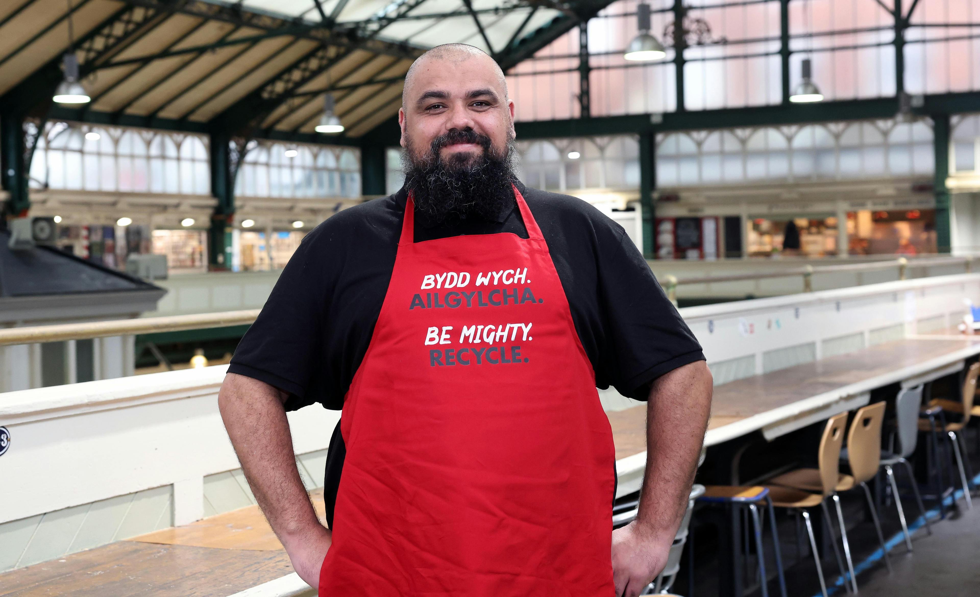 Chef Chris Roberts standing in Cardiff Market wearing a red apron with his hands on his hips.