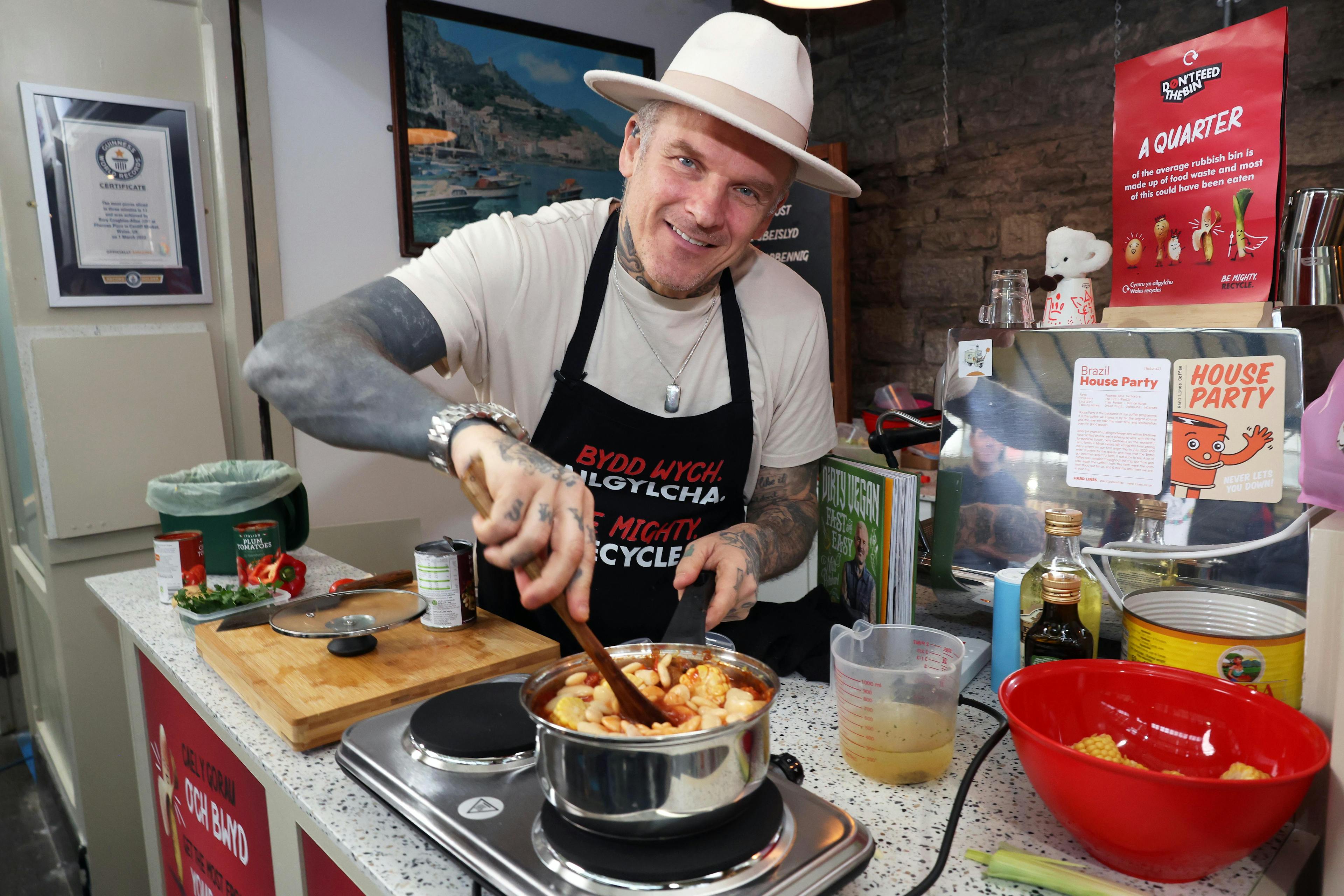 The chef Matt Pritchard standing in Cardiff Market wearing a wide-brimmed hat and string a saucepan of food on a hob.